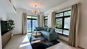 Un salon moderne avec un canapé, une table basse et un téléviseur. De grandes fenêtres, ornées de rideaux, offrent une vue sur la verdure environnante.