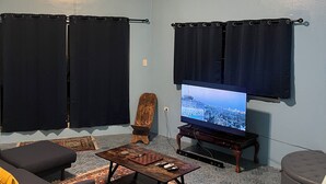 Een woonkamer met een televisie, zwarte gordijnen, een houten stoel, een salontafel en een kamerbreed tapijt met een patroon.