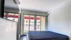A small, narrow room with a bed, a sliding glass door leading to a balcony, and a visible outdoor area with buildings.