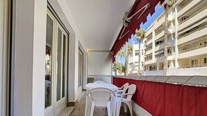 A balcony with a white table and chairs, a red awning, and a view of a multi-story building with balconies.