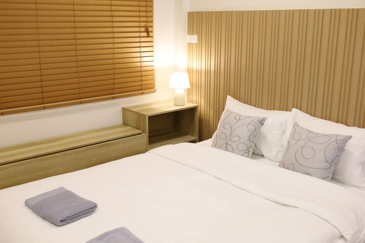 Une chambre d’hôtel avec un lit, une table de chevet avec une lampe, une fenêtre avec des stores et une tête de lit en bois.