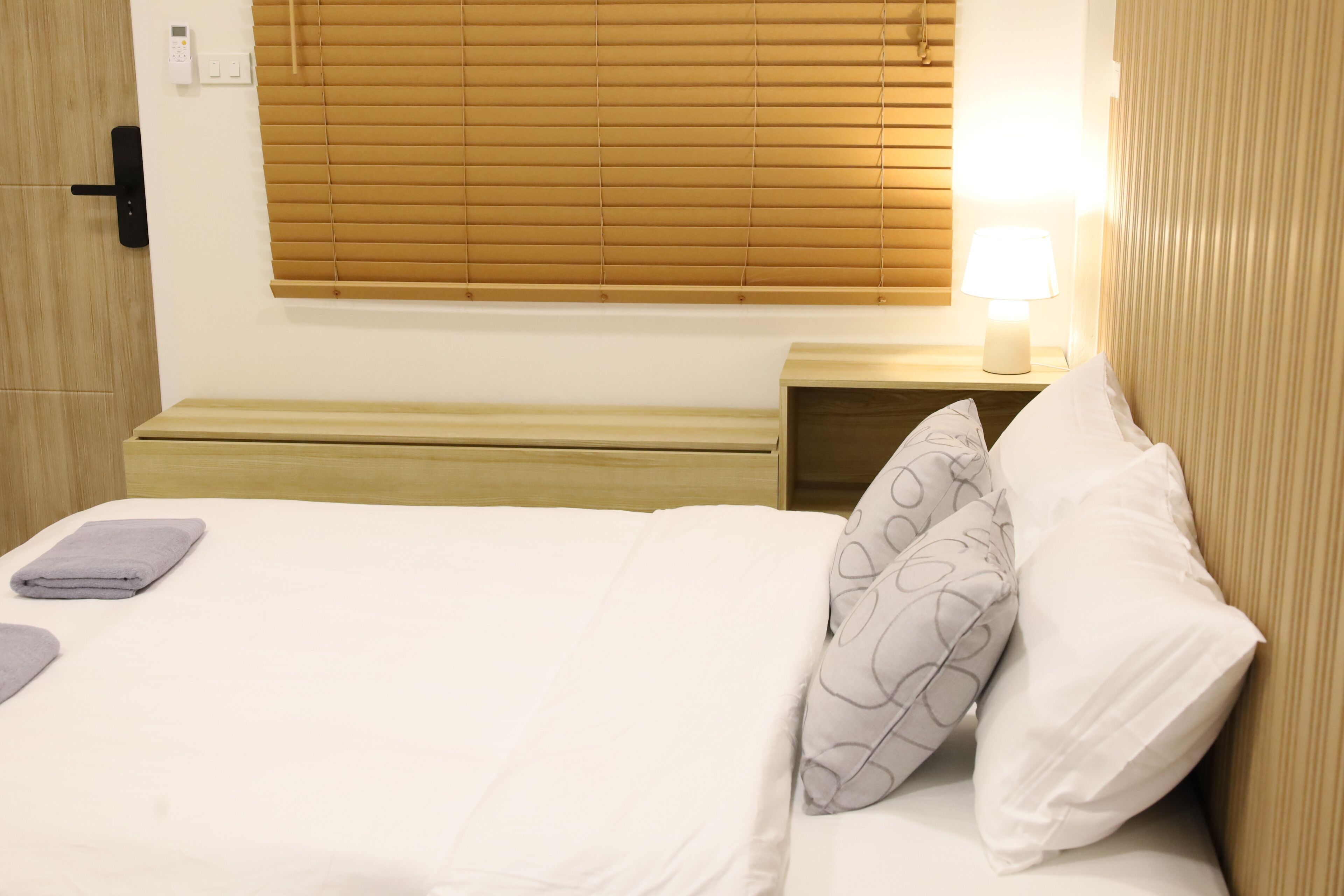 A neatly made bed with white and grey patterned pillows, a bedside table with a lamp, and a window with blinds.