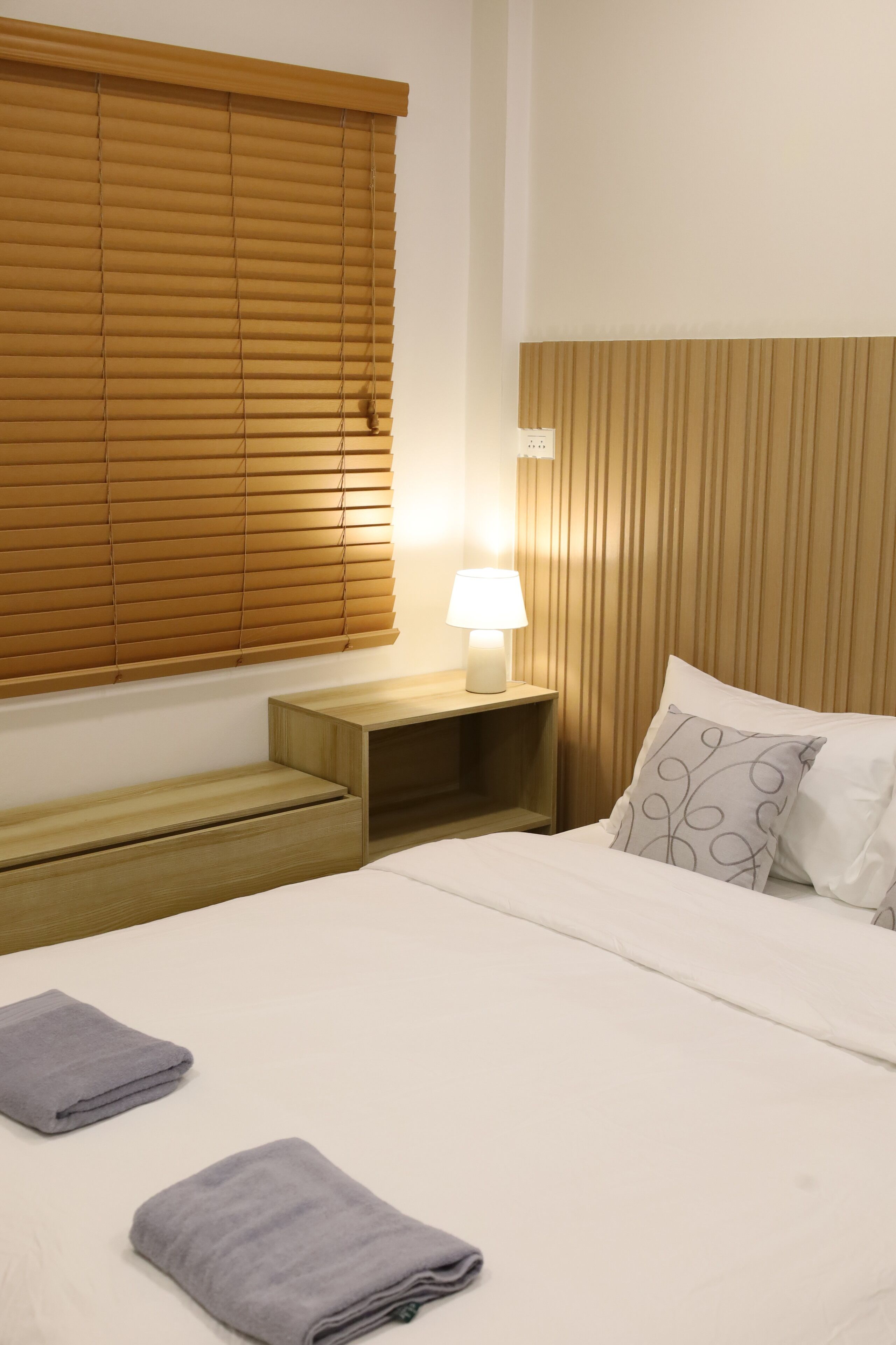 A hotel room with a bed, a bedside table with a lamp, and a window with blinds.