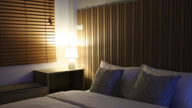 Una cama bien hecha con una colcha blanca y dos almohadas, una mesita de noche con una lámpara, y una pared con persianas verticales.