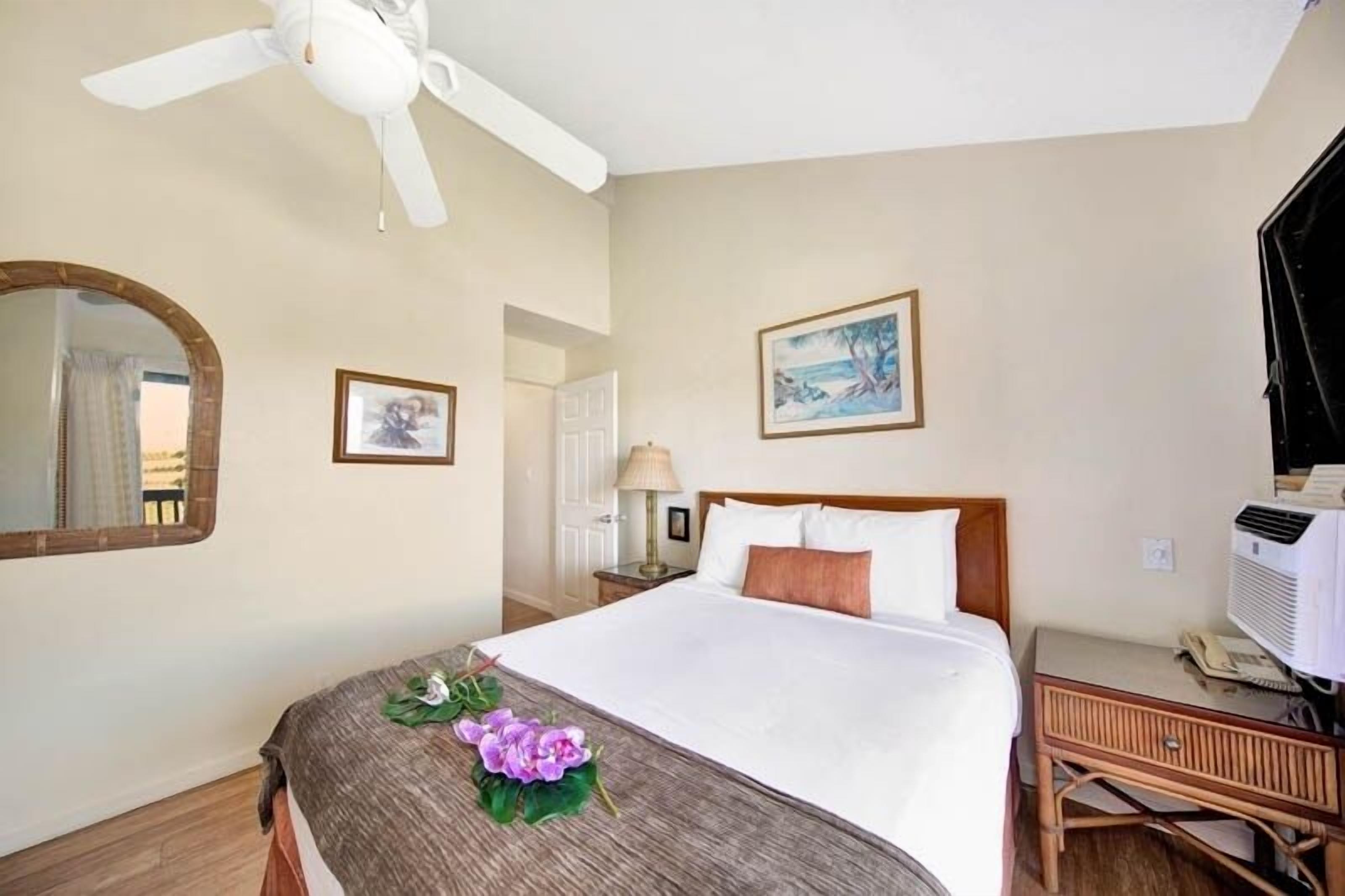 Une chambre d’hôtel avec un lit, un ventilateur de plafond, une table de chevet avec une lampe, un tableau encadré au mur et une fenêtre avec des rideaux.