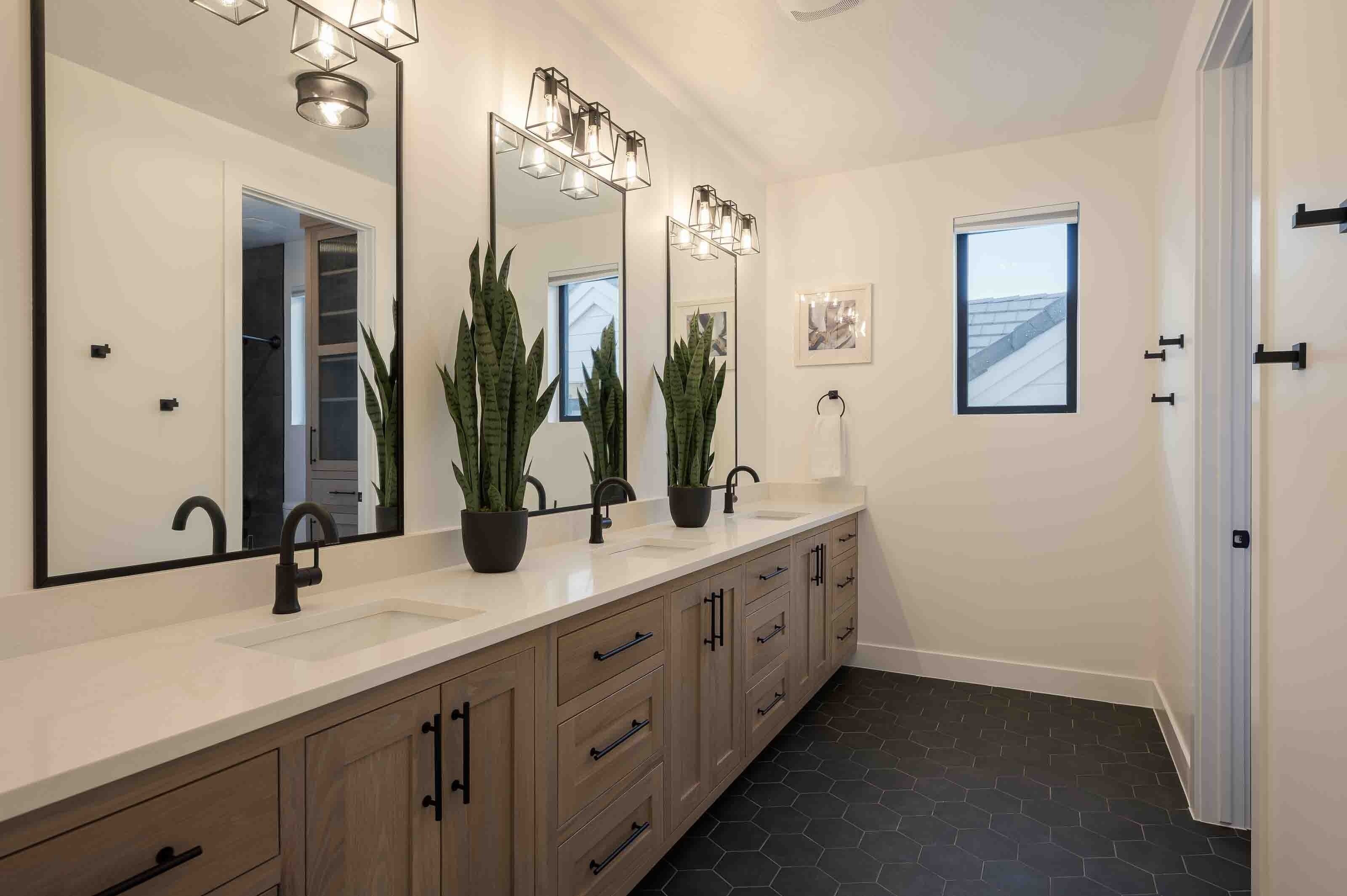 Une salle de bain moderne avec un grand miroir, deux lavabos, des plantes en pot et une fenêtre.