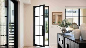 A modern interior with a sliding glass door leading to a balcony, a dark wooden cabinet with decorative items, and a large mirror reflecting the room. - Designer Digs 9 Bedroom Home (St. George)