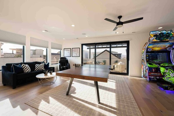 A modern living room with a ping pong table, a black sofa with checkered cushions, a ceiling fan, and a large window with a view of a house.
