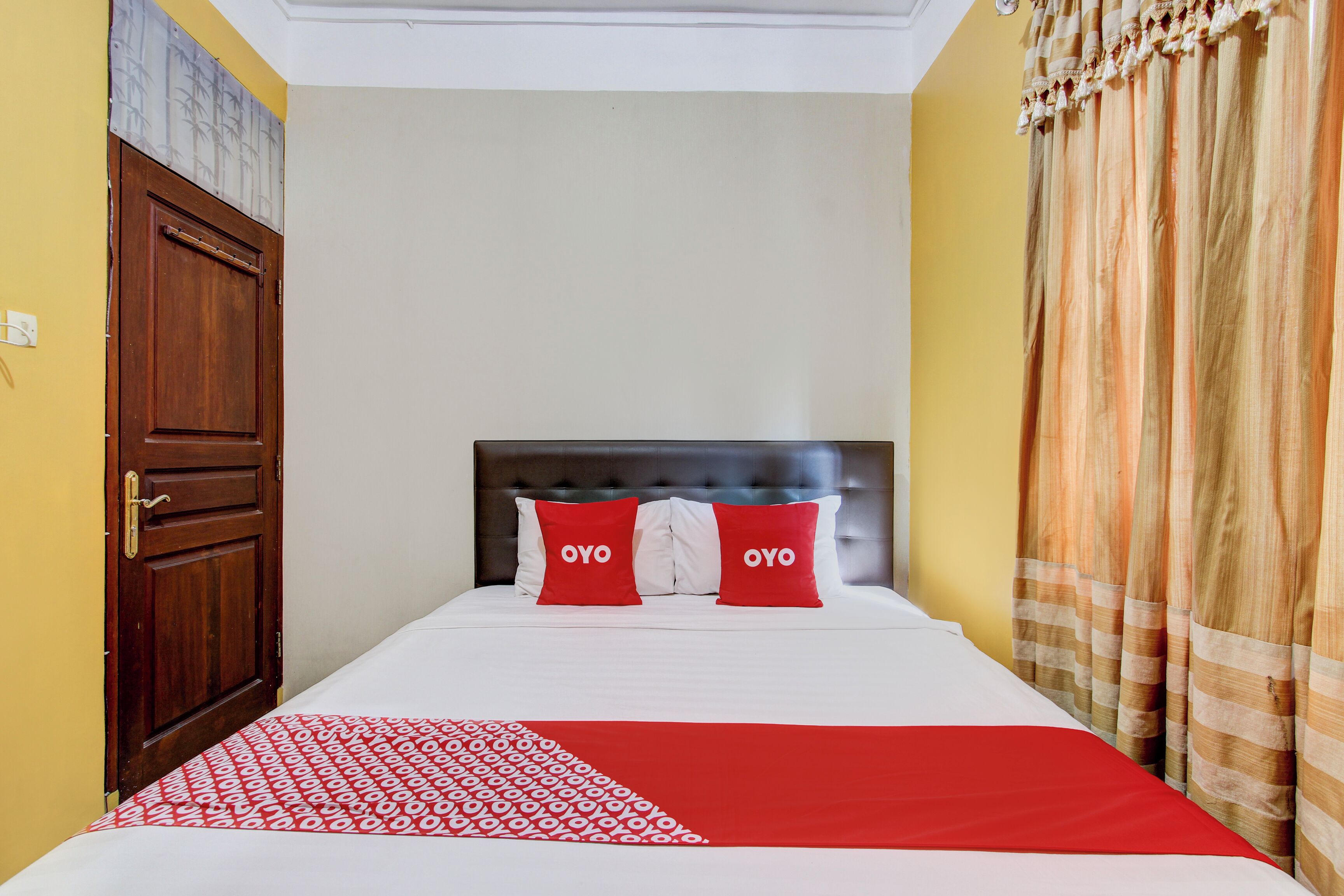 A hotel room with a bed, two red pillows, a patterned throw, a wooden door, and curtains.