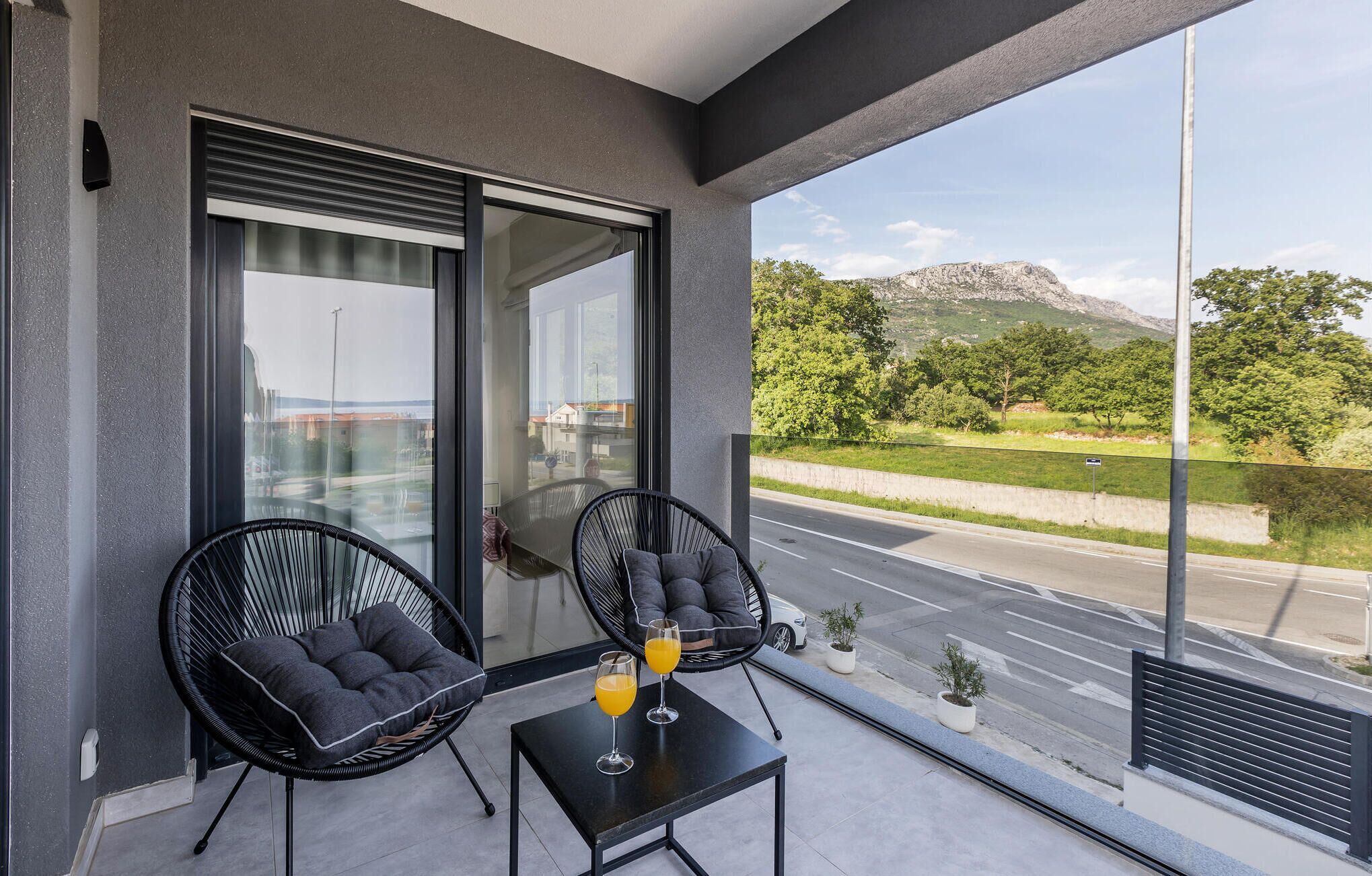 Een balkon met twee zwarte stoelen en een kleine tafel, twee glazen sinaasappelsap, en uitzicht op een weg en begroeiing.