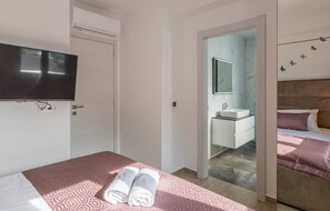 Une chambre d’hôtel comprenant un lit, une télévision, une salle de bain avec un miroir et un lavabo, ainsi qu’un couloir menant à une autre chambre.