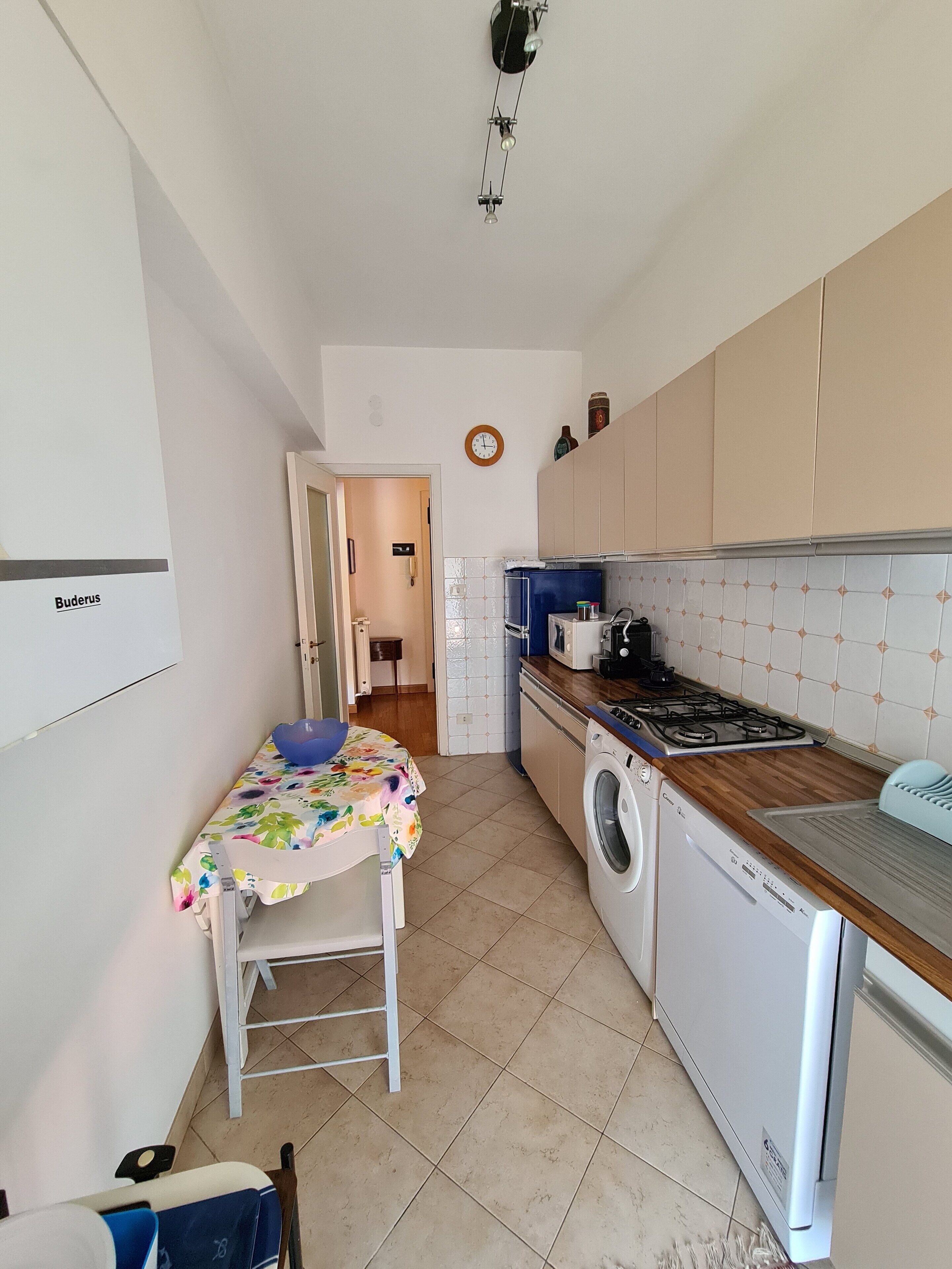 A compact kitchen with a washing machine, a stove, and a sink. There's a small table with a floral tablecloth and a chair.