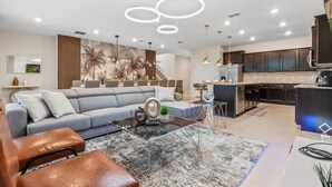 A modern living room with a sofa, coffee table, and ottoman. There is a kitchen area with dark cabinets and a large island.
