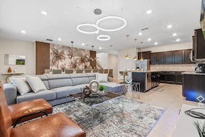 A modern living room with a sofa, coffee table, and ottoman. There is a kitchen area with dark cabinets and a large island. - 3508 Lavender Windsor · New Luxury Home | Windsor Island by Disney w/Pool (Davenport)