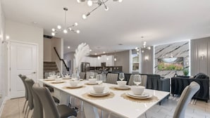 Un espace de repas moderne avec une longue table dressée pour six personnes, une cuisine en arrière-plan et un coin salon avec un canapé et un téléviseur.