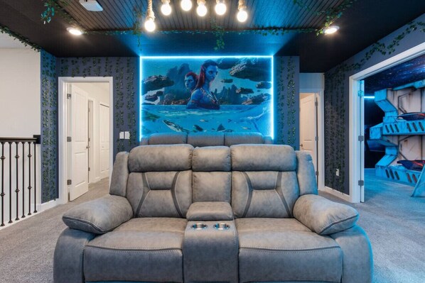 A home theater room with a large reclining sofa, a wall-mounted screen displaying a movie scene, and a staircase leading to another level.