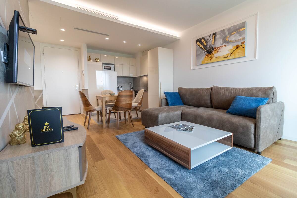 A modern living room with a grey sofa, a coffee table, and a TV mounted on the wall. There is a dining area in the background with a table and chairs.