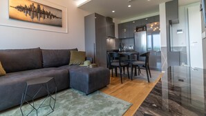 Un salon moderne avec un canapé d’angle, un coin repas avec des chaises et une cuisine dotée de meubles sombres et d’appareils électroménagers en acier inoxydable.