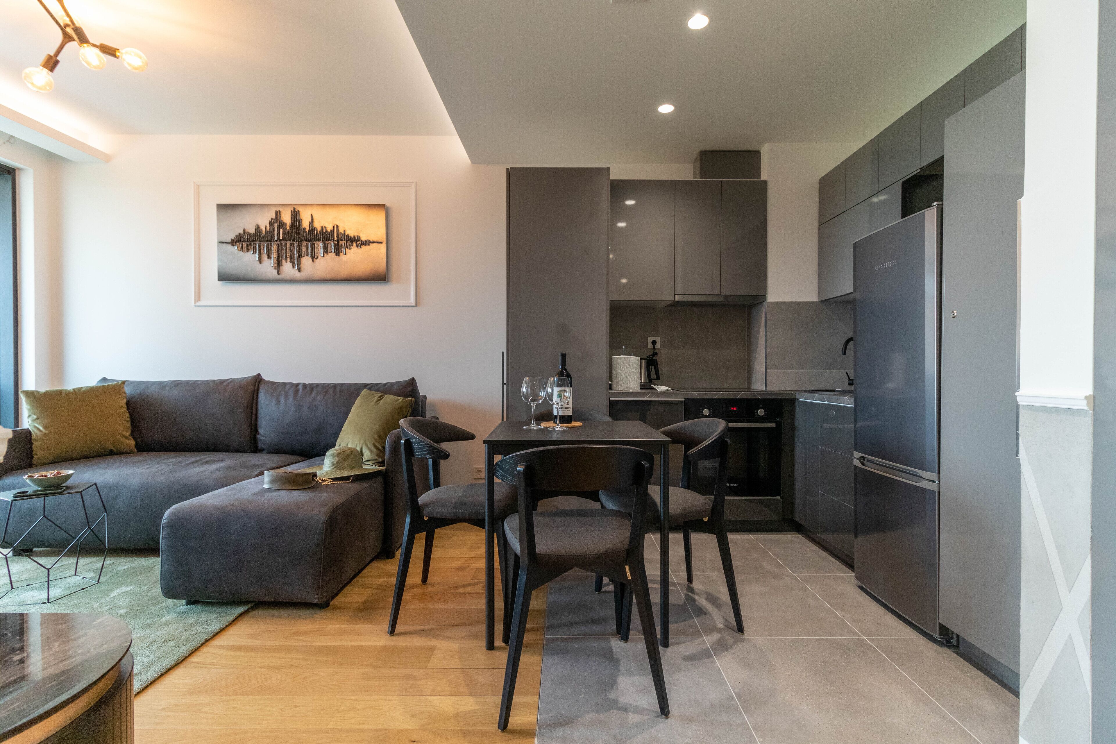 A modern living room with a sectional sofa, a dining table with chairs, and a kitchen area with a refrigerator and cabinets.