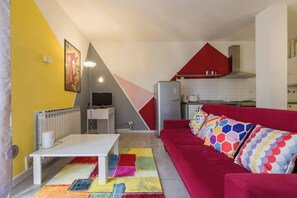 Un espace de vie compact comprenant un coin cuisine, un canapé rouge agrémenté de coussins à motifs, une table blanche et un tapis coloré.