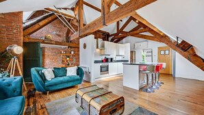 A loft-style apartment with exposed wooden beams, a kitchen, and a living area with a blue sofa and a vintage trunk.