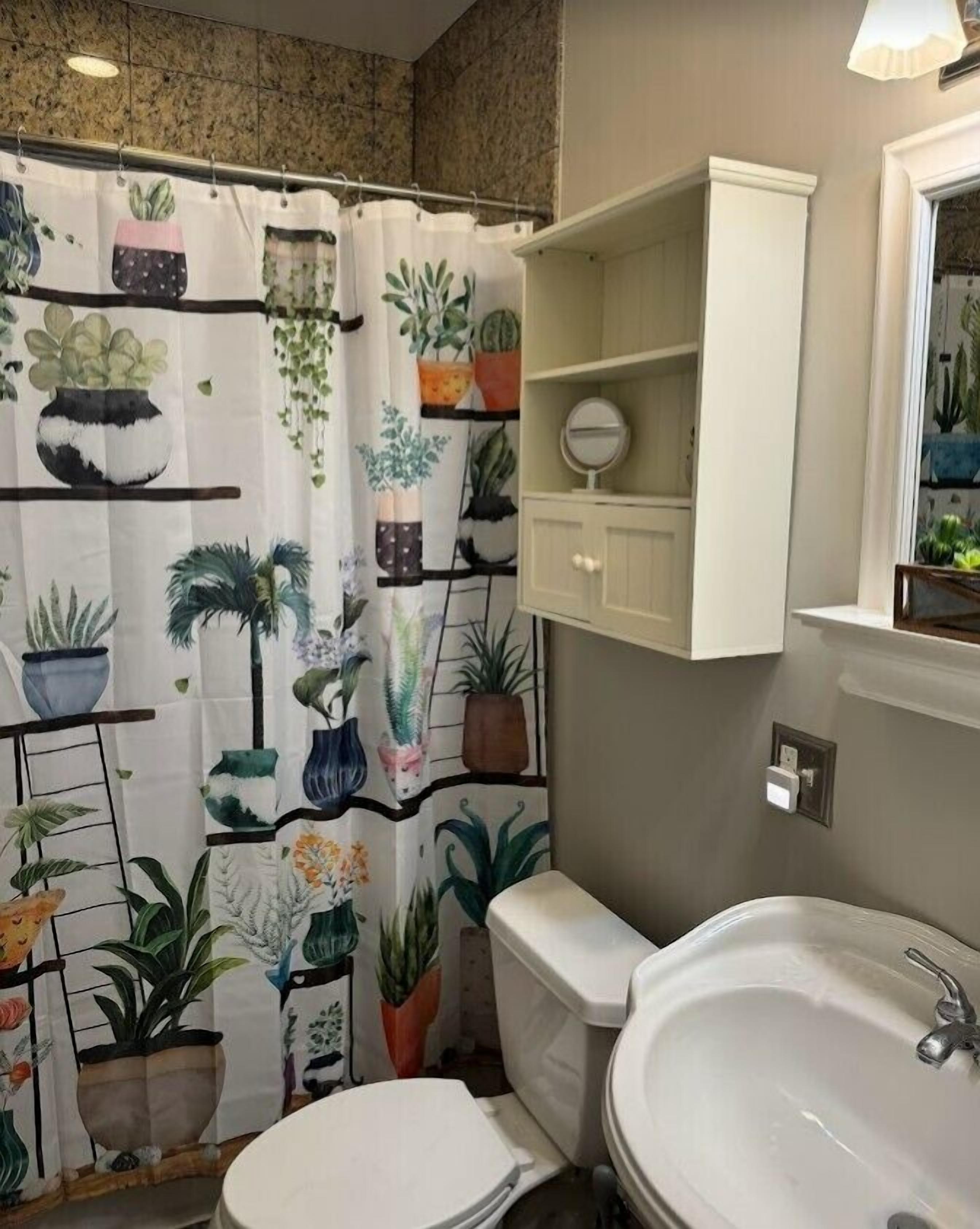 Ein Badezimmer mit Duschvorhang mit Pflanzenmuster, Toilette, Waschbecken und einem kleinen Schrank mit Spiegel.