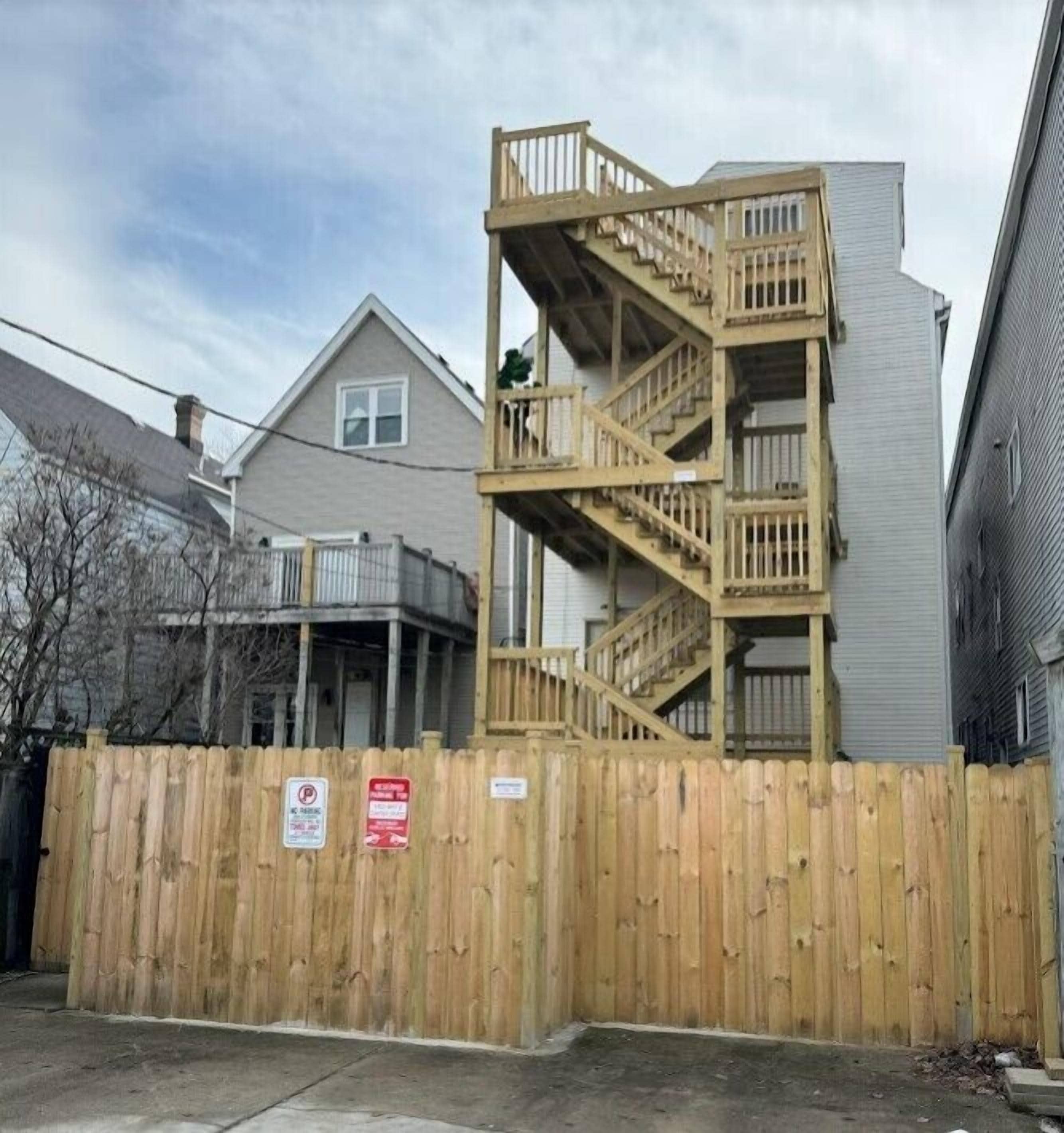 Un edificio residencial con una escalera de madera adosada al lateral, una valla de madera en la parte frontal y un cielo despejado.