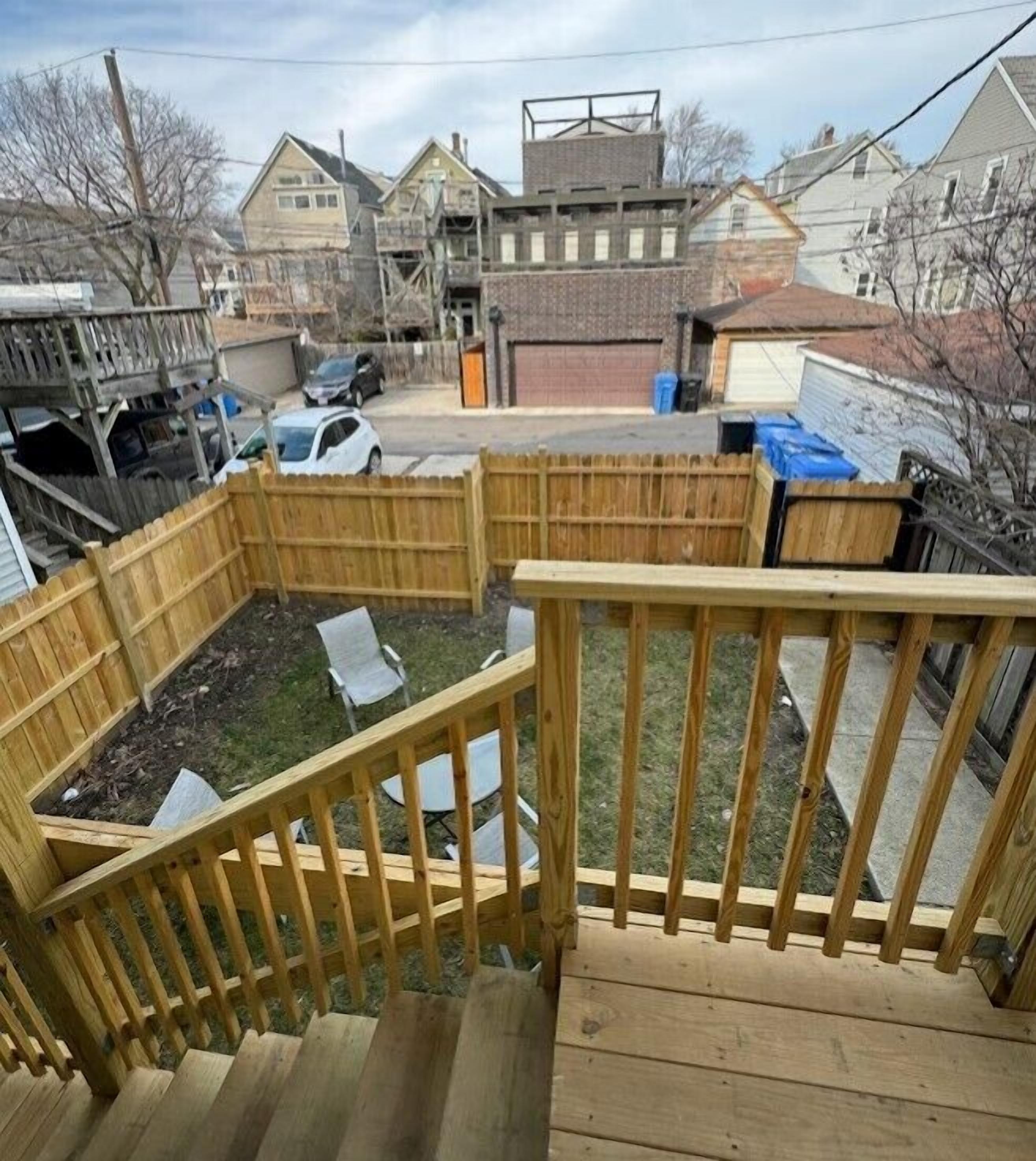 A wooden deck with a wooden railing, surrounded by a fenced yard with chairs and a small pool.