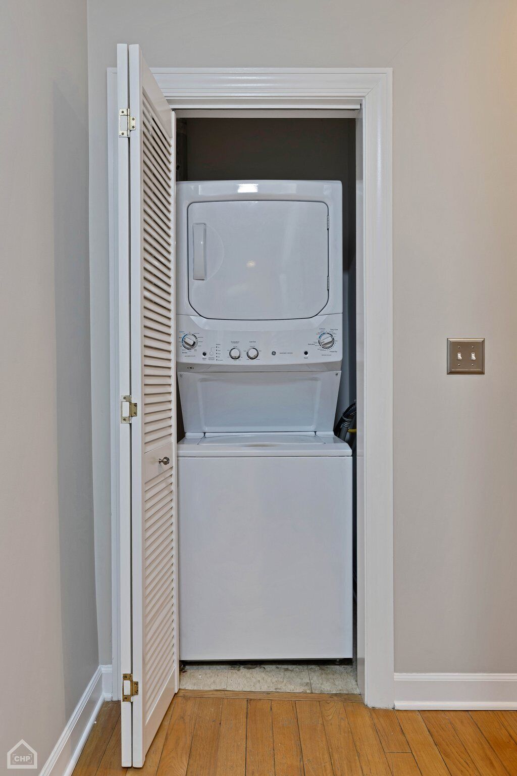 A compact laundry room with a front-loading washer and dryer, a door with a latch, and a light switch on the wall.