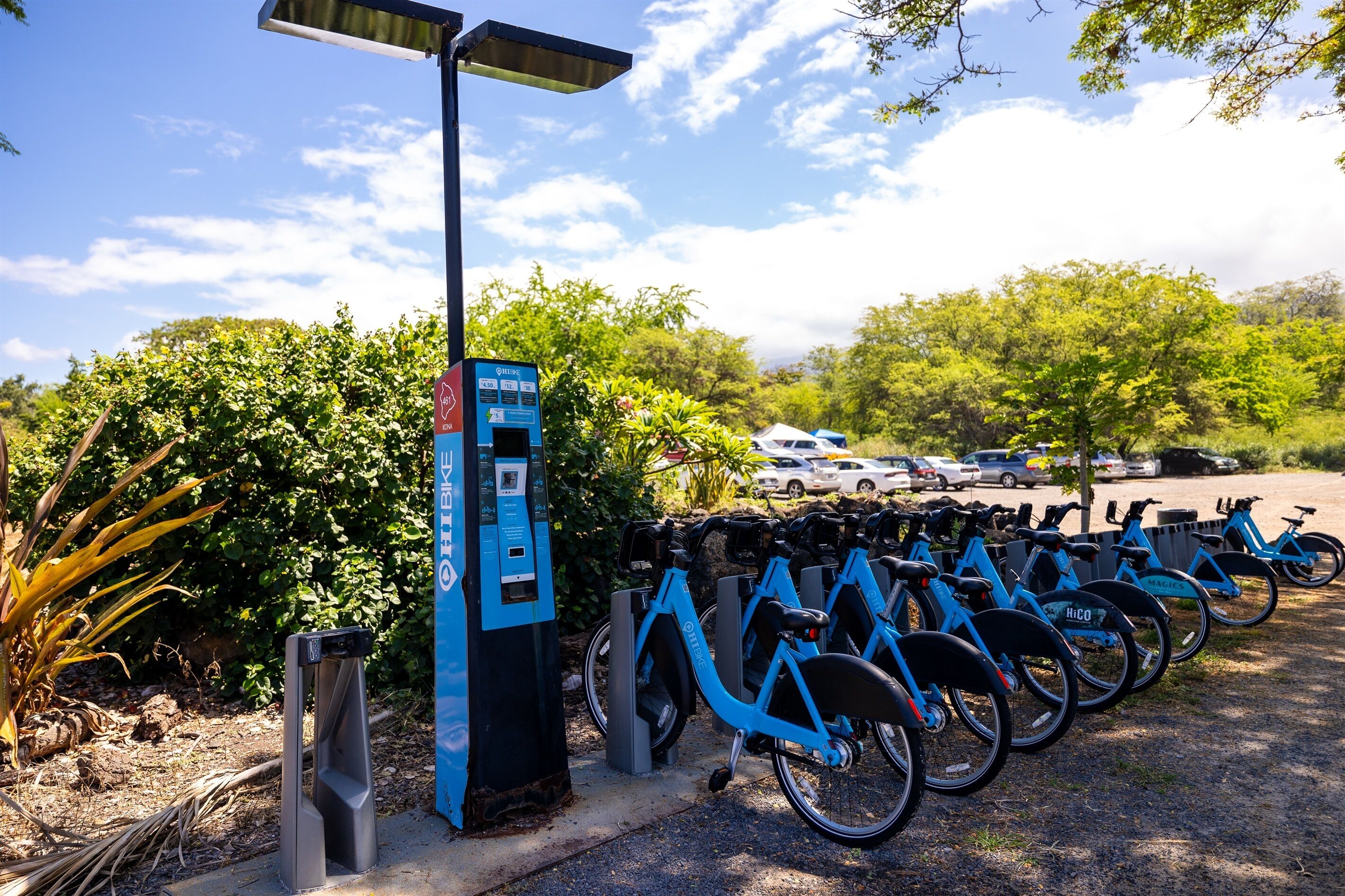 A row of blue bicycles parked next to a solar-powered charging station in a parking lot with trees and cars in the background.