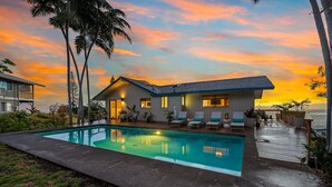 A modern house with a swimming pool, a deck with lounge chairs, and a view of the ocean at sunset.