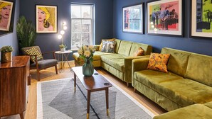 Ein modernes Wohnzimmer mit einem grünen Sofa, einem Holztisch und einer Topfpflanze. An den Wänden befinden sich gerahmte Kunstwerke und ein Fenster mit Tageslicht.