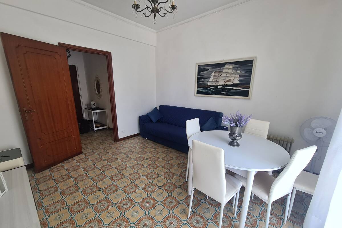 A room with a dining table, chairs, a blue sofa, a white fan, and a ship painting on the wall.