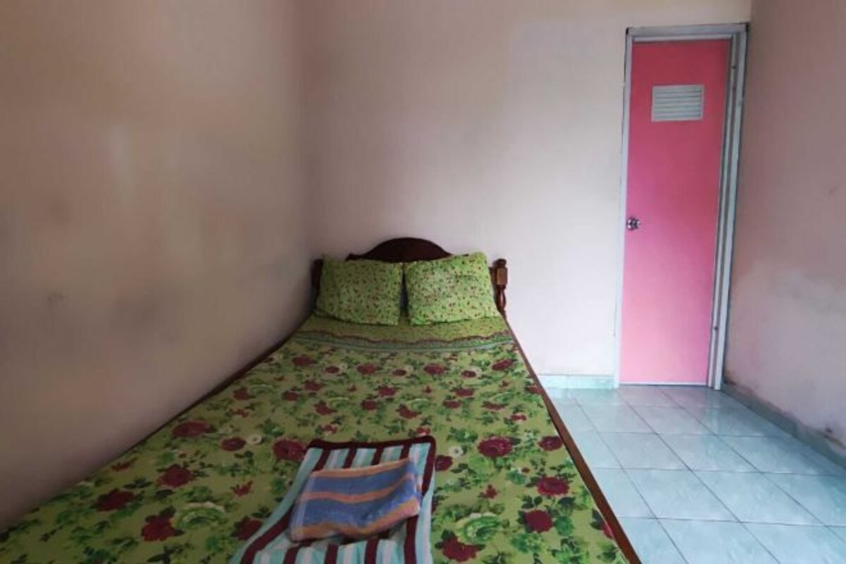 Una habitación pequeña con una cama individual, una colcha de estampado floral y almohadas, una manta a rayas en la cama, y una puerta rosa al fondo.