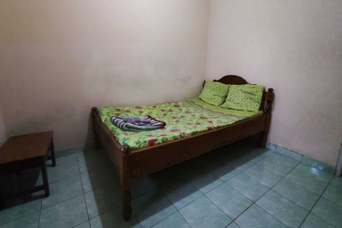 A small, single-bed room with a wooden frame, green floral bedding, and a small wooden stool.