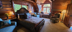 A log cabin bedroom with a large bed, two chairs, a small table, and a window with a view of trees.