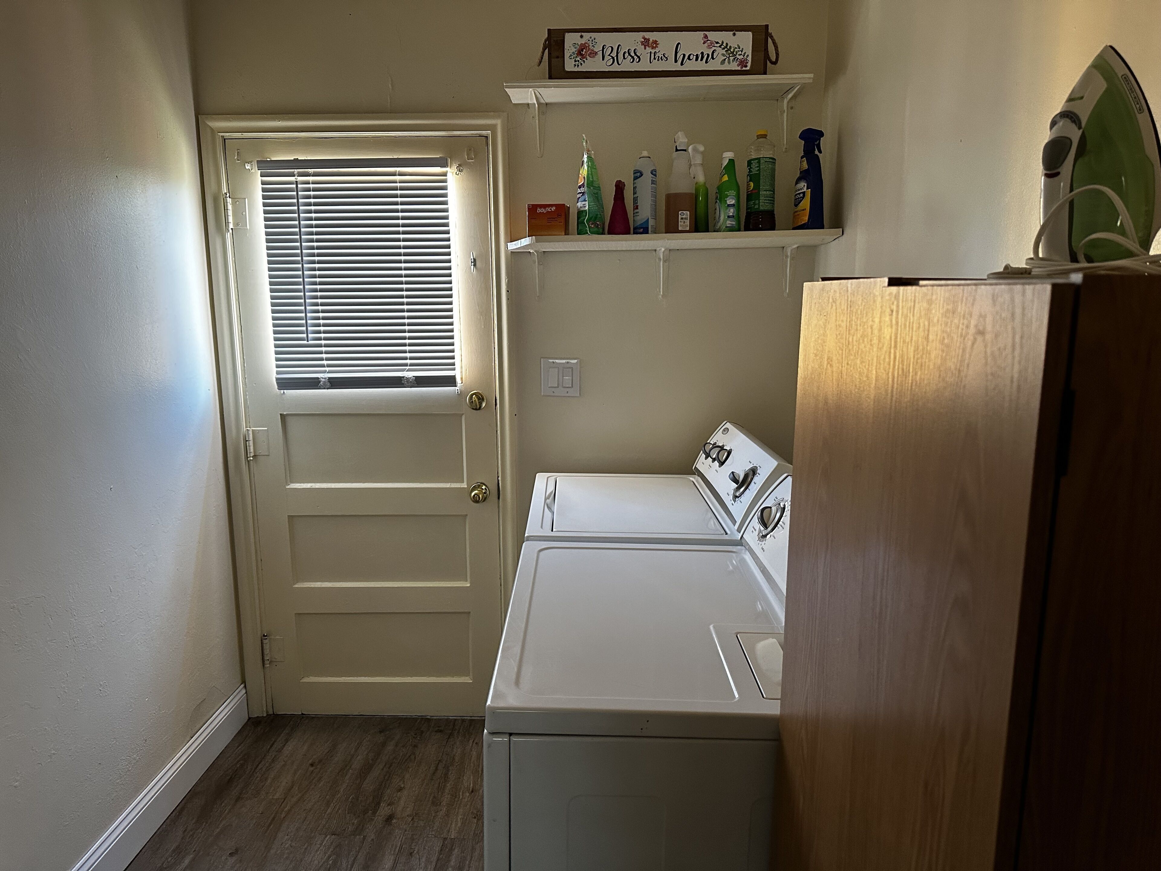 A laundry room with a white washer and dryer, a door with a window, and a shelf with cleaning supplies.
