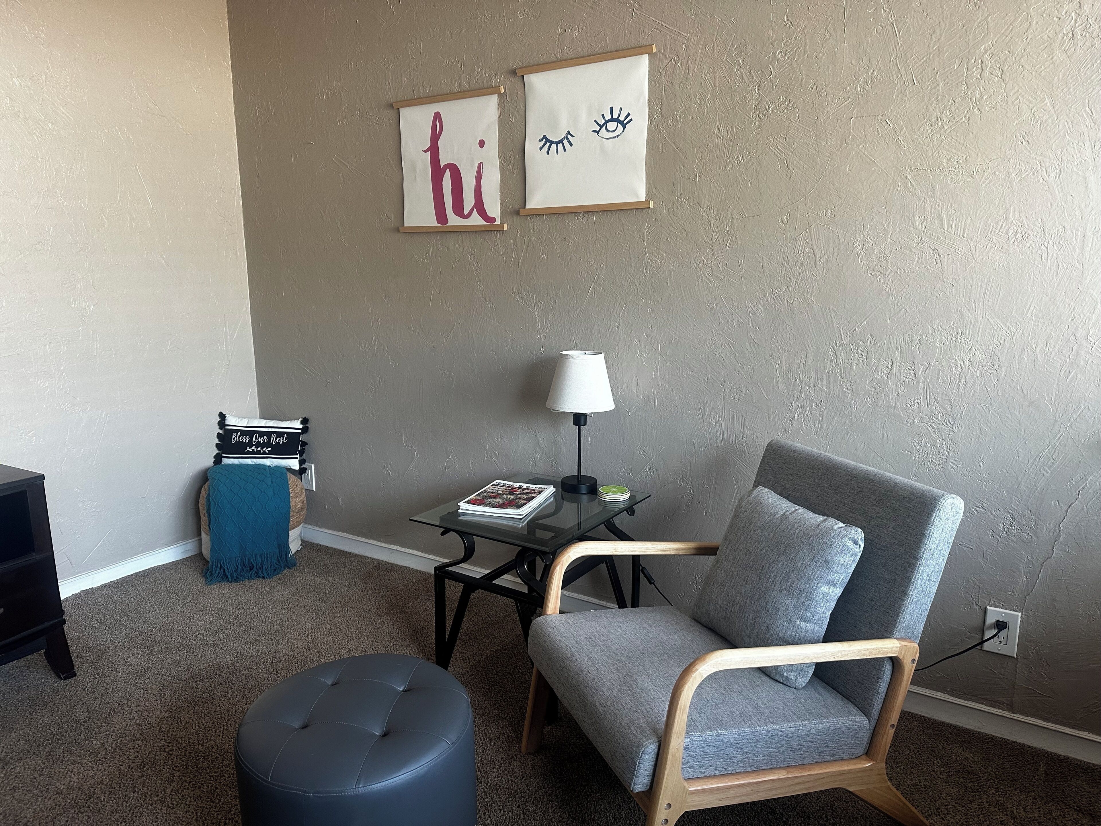 A living room with a grey armchair, a round ottoman, a small table with a lamp, and two framed pictures on the wall.