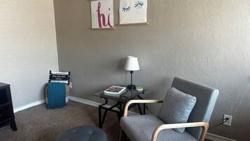 A living room with a grey armchair, a round ottoman, a small table with a lamp, and two framed pictures on the wall.