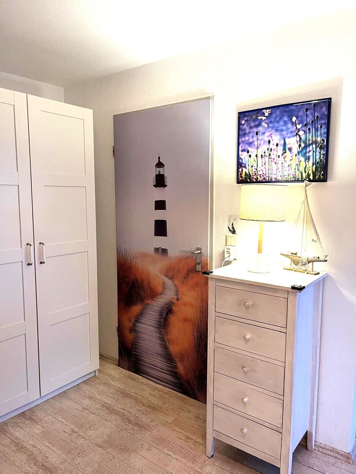 Une pièce avec une armoire blanche, un bureau avec des tiroirs, une lampe et un mur orné d’un tableau.