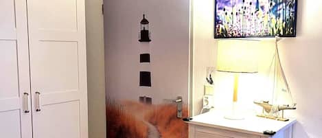 A room with a white wardrobe, a desk with drawers, a lamp, and a wall with a painting.