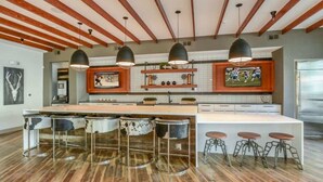 Uma cozinha moderna com vigas de madeira, luminárias pendentes, uma ilha central com banquetas e uma televisão grande.