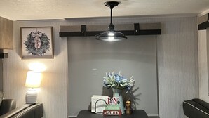 A dining area with a table set for two, a wall-mounted light, a decorative wreath, and a ceiling fan. - Cozy camper in Clemson Hills.  5 minutes from Clemson University.  Pet friendly! (Seneca)