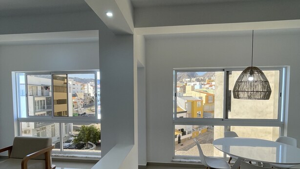 A modern living room with a dining area, large windows offering city views, and a hanging light fixture. - Biss Apartments Mindelo - T1 Madalena nearby the beach (Mindelo)