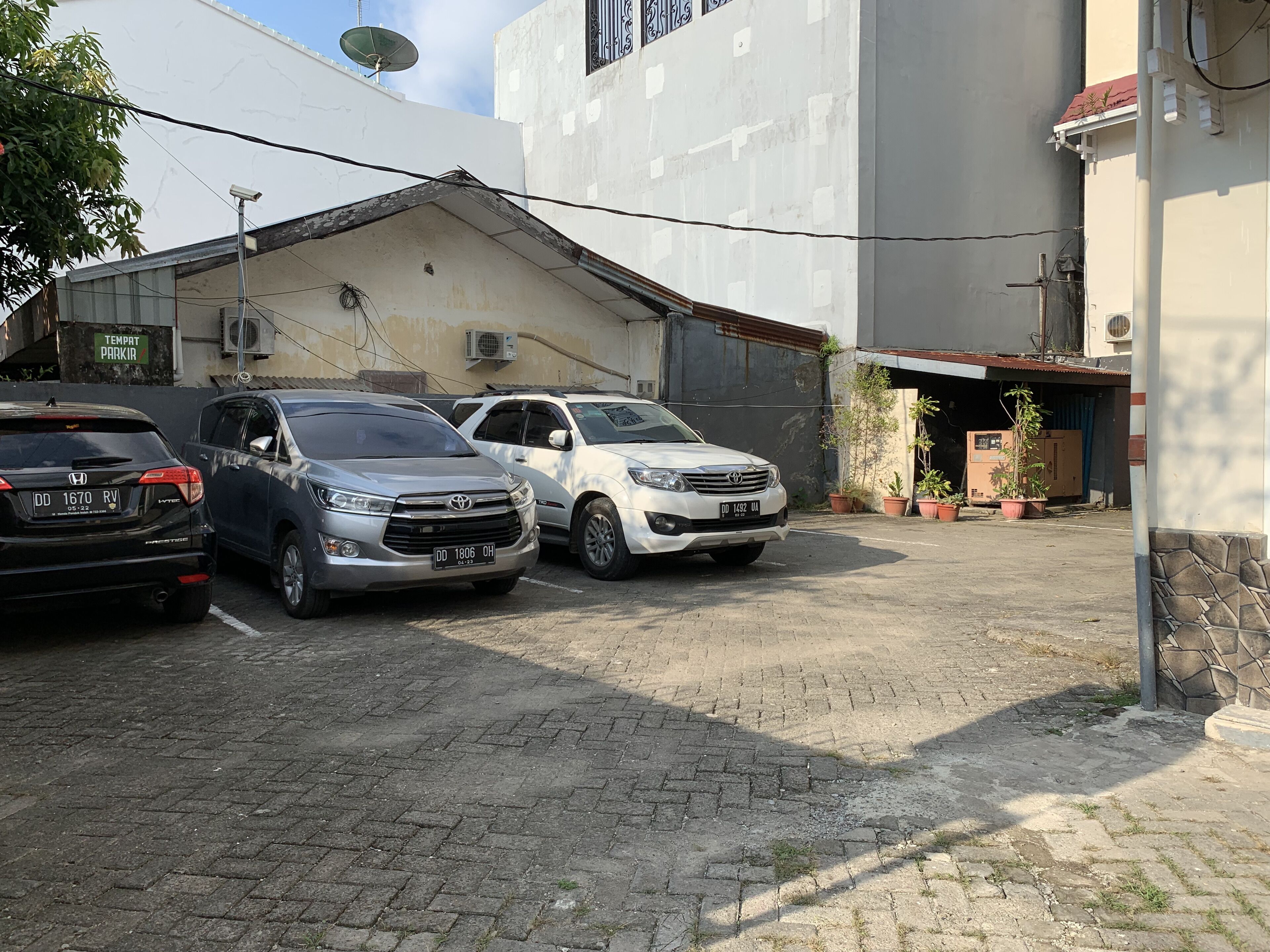 A parking area with three cars parked in front of a building with a satellite dish and air conditioning units.
