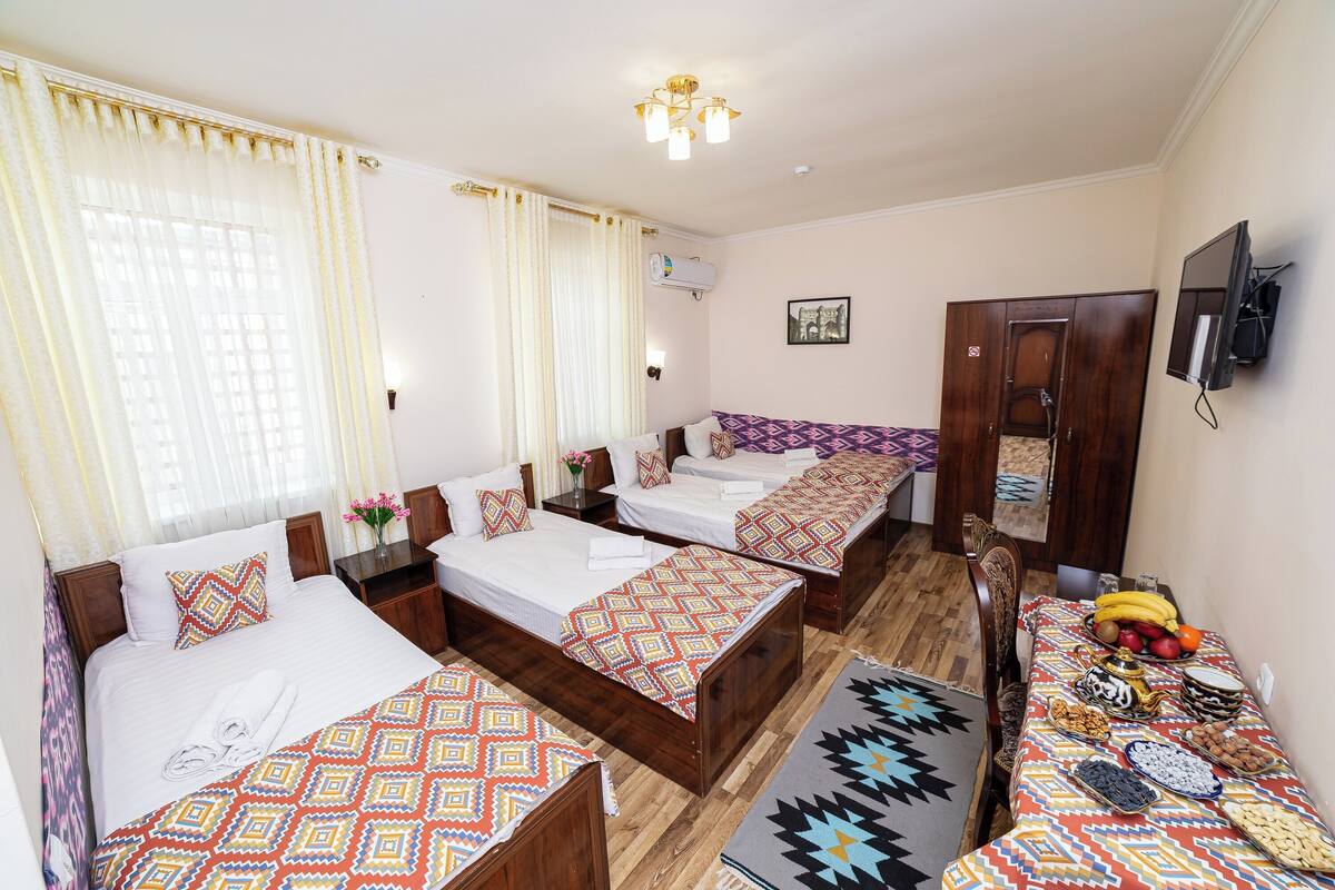 A hotel room with three beds, a dining table with fruits and nuts, a TV, and a wardrobe.
