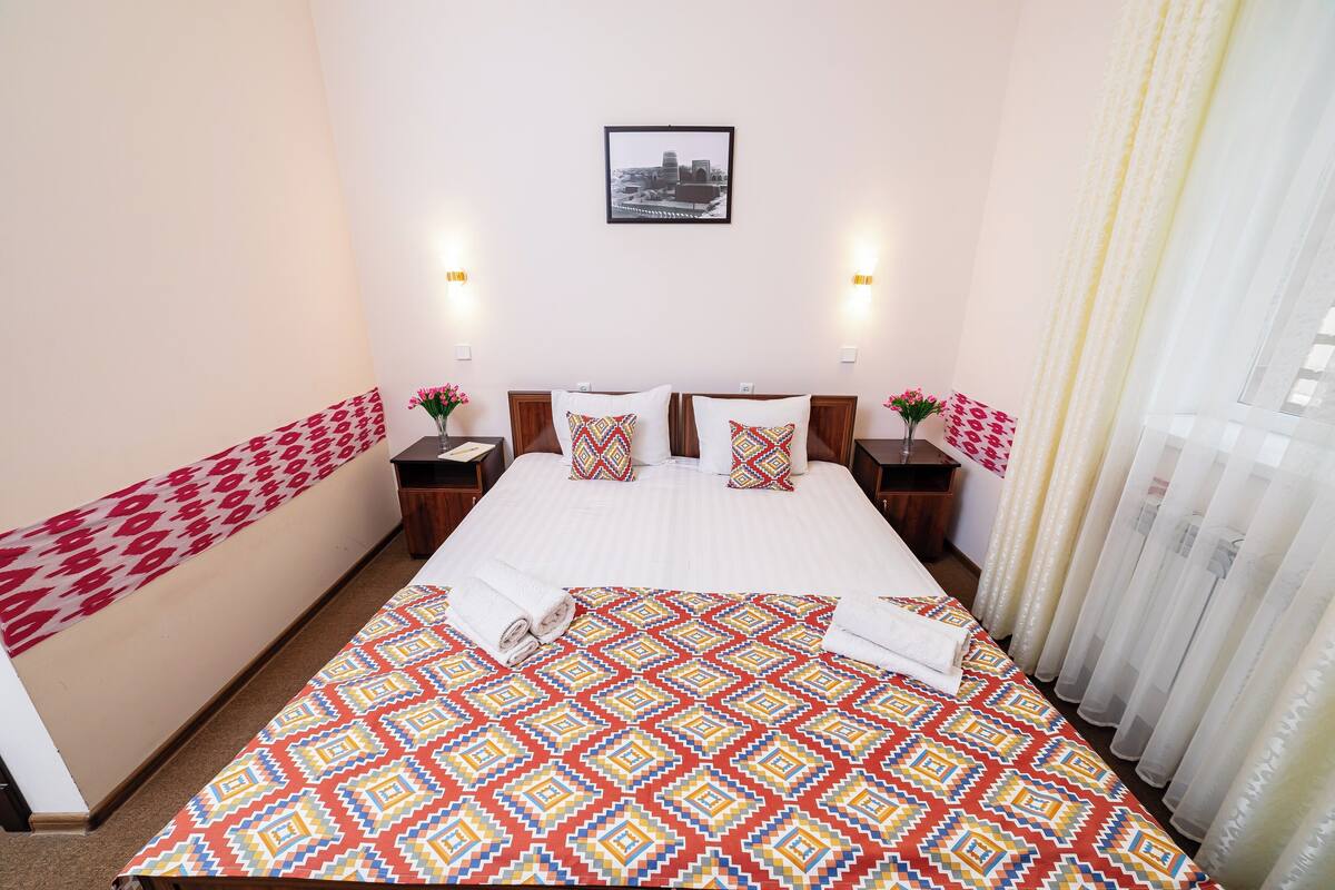 A hotel room with a large bed, two bedside tables with lamps, a framed picture on the wall, and a window with sheer curtains.