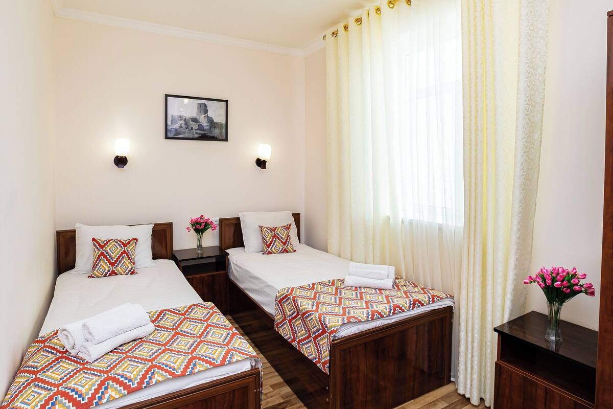 A hotel room with two beds, a wooden floor, a patterned rug, a nightstand with a vase of flowers, and a window with curtains.