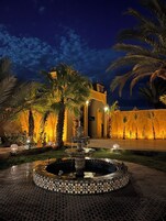 Vista nocturna de un patio con una fuente, palmeras y un edificio con iluminación cálida.