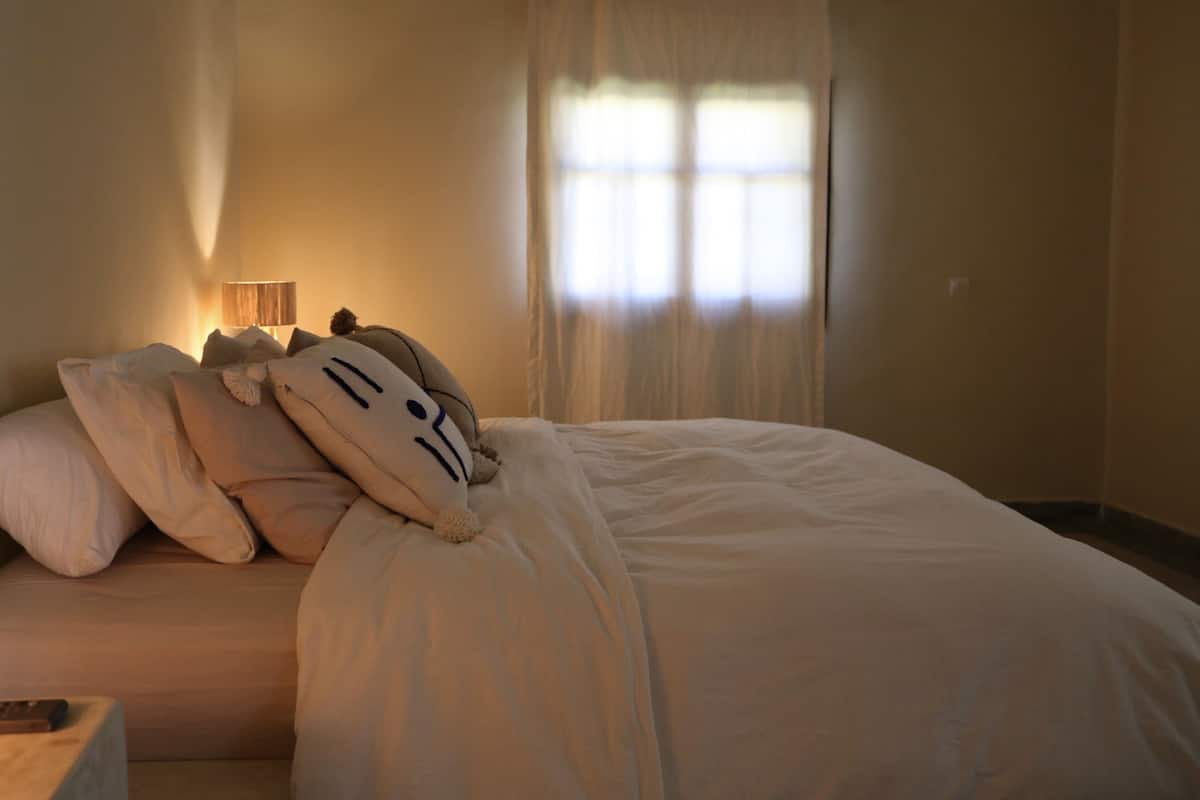 A neatly made bed with white bedding and decorative pillows, a bedside lamp, and a remote control on a wooden nightstand.